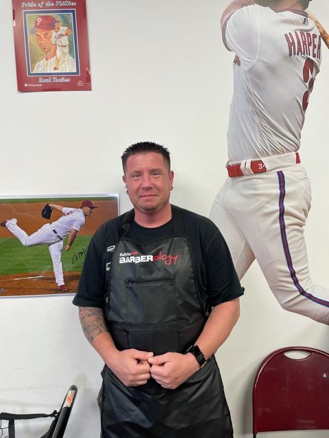 A smiling barber in a salon, wearing an apron with scissors, baseball picture in background.